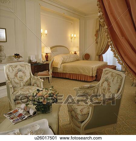 Stock Image - 18th century-style armchairs in large townhouse bedroom with lighted wall-lights above bed with beige silk bedcover. Fotosearch