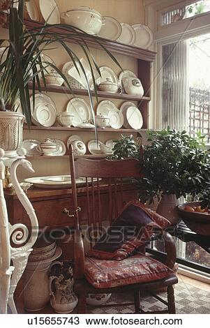 Stock Image - Antique chair in front of dresser with white crockery in corner of dining room with houseplant on low table. Fotosearch