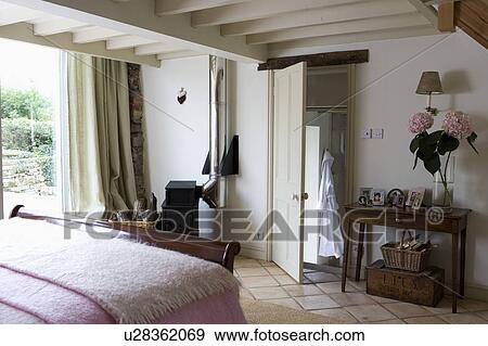 Antique Console Table Beside Open Door In Cottage Bedroom With White Ceiling Beams And Cream Curtains Stock Photo