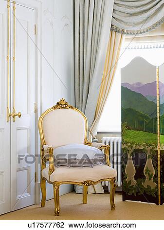 Stock Image - Antique gilt chair with cream upholstery in front of window with grey swagged curtain. Fotosearch