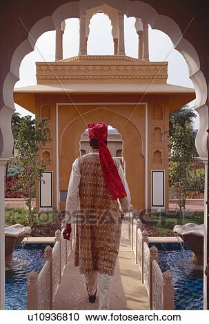 Stock Image - Back view of man in traditional Rajastani clothes crossing footbridge over water at Rajvilas Hotel. Fotosearch