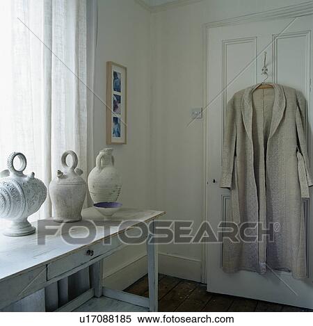 Stock Photography - Beige linen coat on door in small hall with collection of Greek ceramic pots on pale grey wooden console table. Fotosearch