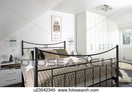 Stock Photography - Beige textured throw on black metal bed in modern white attic bedroom with white fitted wardrobe. Fotosearch