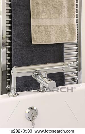 Stock Image - Beige towel on chrome heated towel rail above chrome tap on bath. Fotosearch