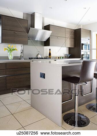 Stock Photograph - Black leather and chrome stools at breakfast bar on island unit in modern kitchen with dark wood fitted units. Fotosearch