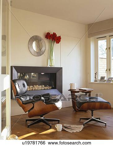 Stock Photograph - Black leather Charles Eames armchair and stool in front of modern fireplace in modern living room. Fotosearch