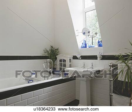 Stock Image - Black tiled dado and matching tiled bath edging in modern white bathroom with white pedestal basin. Fotosearch
