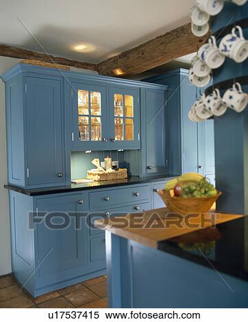 Stock Photography - Blue fitted cupboards with lighted shelves behind glazed cupboard doors in traditional country kitchen. Fotosearch