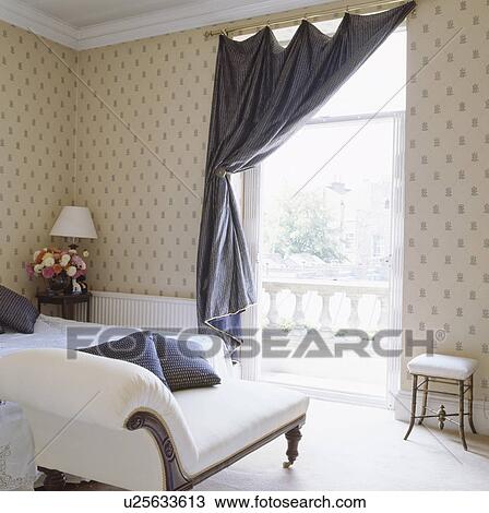 Stock Image - Blue silk curtain on tall window in traditional bedroom with white antique chaiselongue and patterned wallpaper. Fotosearch