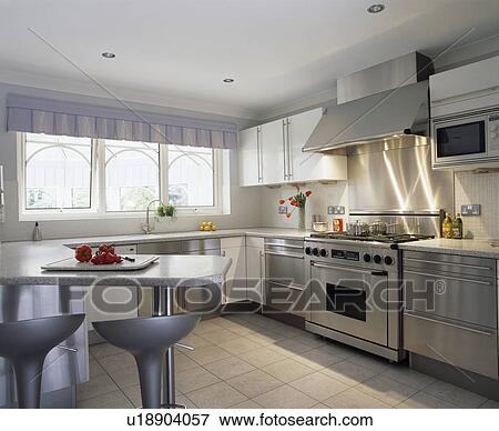 Stock Photo - Bomba stools at breakfast bar in modern kitchen with stainless steel fitted units and range oven. Fotosearch