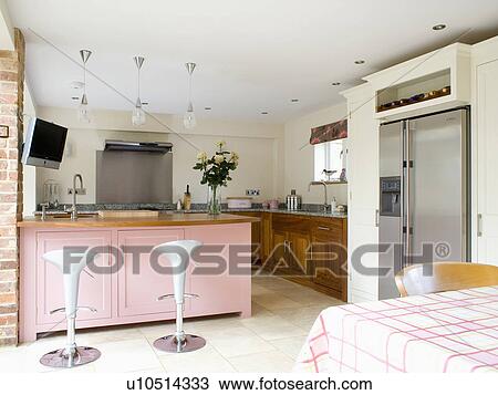 Stock Image - Bomba stools at breakfast bar on pastel pink peninsular unit in modern cream kitchen with stainless steel fridge-freezer. Fotosearch