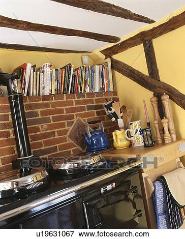 Stock Photo - Books on shelf above exposed brick wall and black Aga oven in yellow cottage kitchen with wall beams. Fotosearch