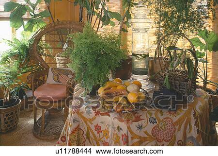 Picture - Bowl of fruit and lush green houseplants on table with patterned cloth beside cane peacock chair. Fotosearch