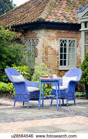 Bright blue painted Lloyd Loom armchairs and table on paved patio in front of country house with green Gothic windows View Large Photo Image Stock Photograph - Bright blue painted Lloyd Loom armchairs and table on paved patio in front of country house with green Gothic windows. Fotosearch
