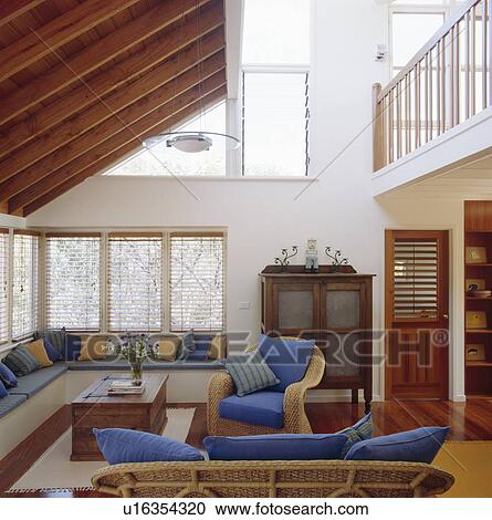 Stock Image - Built-in seating below windows in double-height living room with wicker armchair. Fotosearch