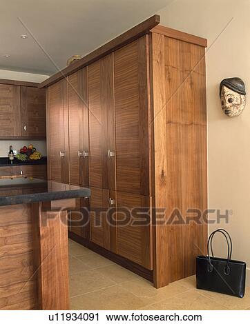 Stock Image - Ceramic mask on wall beside fitted cherrywood storage cupboard in modern kitchen. Fotosearch