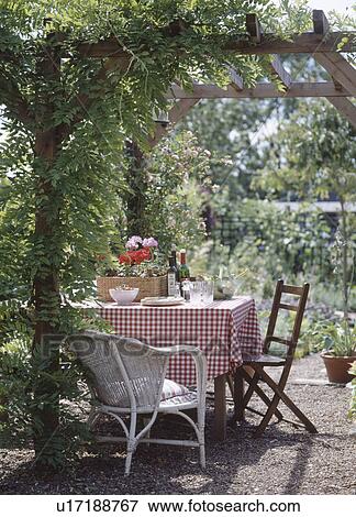 Stock Photo - Chairs and table with red checked cloth below climbing plant on pergola in country garden. Fotosearch