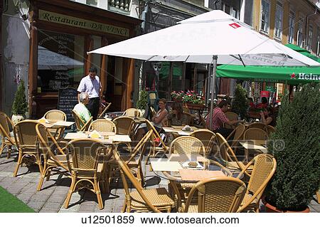 Stock Photo - Chairs and tables below umbrellas on pavement outside traditional Romanian restaurant&#10;. Fotosearch