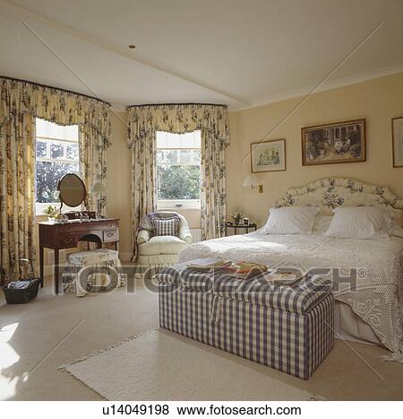 Stock Photo - Checked upholstered ottoman in pastel yellow bedroom with patterned yellow floral curtains and white bedlinen. Fotosearch