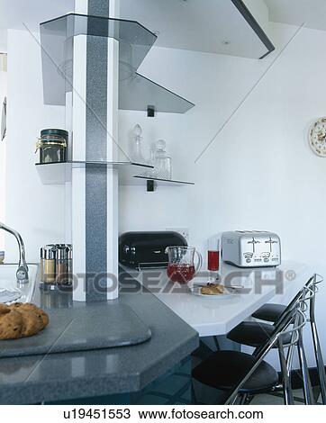 Stock Image - Chrome toaster on triangular breakfas tbar below glass shelves in modern white kitchen. Fotosearch