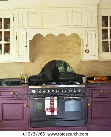 Stock Photograph - Close-up of black range oven in purple fitted unit below fitted cream cupboard in country kitchen. Fotosearch