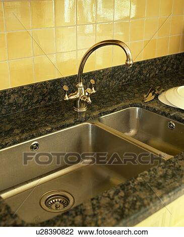 Stock Image - Close-up of chrome tap and stainless-steel double sink below yellow tiled wall. Fotosearch