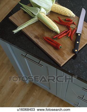 Stock Image - Close-up of corn and chillies with large knife on wooden chopping board. Fotosearch