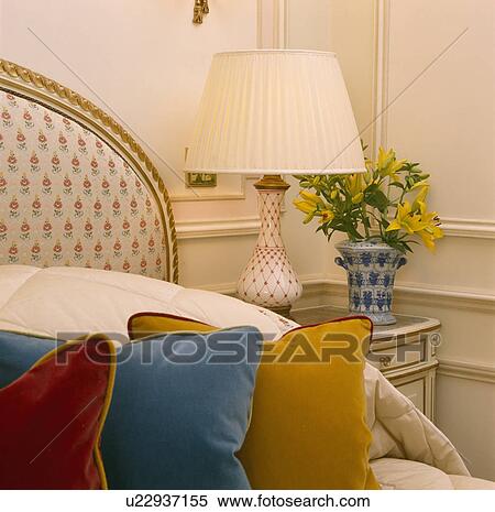 Stock Photography - Close-up of cream lamp on table beside bed with velvet cushions . Fotosearch