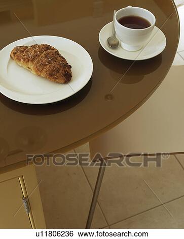 Stock Photograph - Close-up of cup of coffee and croissant on white plate on dark wood table. Fotosearch