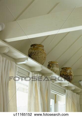 Stock Image - Close-up of earthenware pots on shelf above white curtain. Fotosearch