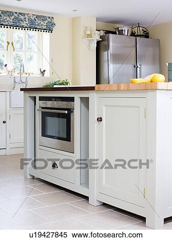 Stock Photography - Close-up of fitted microwave oven in white island unit in cottage kitchen. Fotosearch