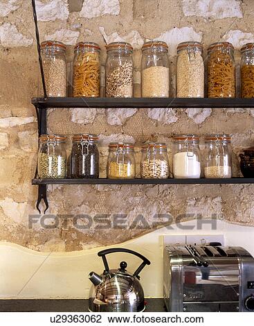Stock Image - Close-up of glass jars on storage shelves on stone wall above chrome kettle and toaster. Fotosearch