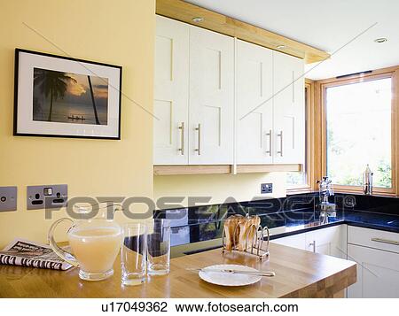 Stock Image - Close-up of glass jug of fruit juice and glasses on kitchen worktop below picture on wall. Fotosearch