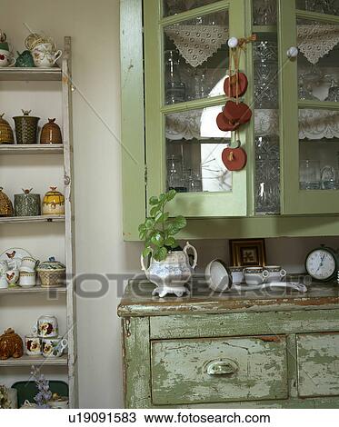 Stock Image - Close-up of green kitchen cupboard with glazed doors above distressed green chest-of-drawers. Fotosearch