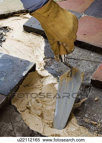 Stock Photography - Close-up of hand wearing rubber glove using trowel to apply cement before laying old terracotta paving tiles. Fotosearch