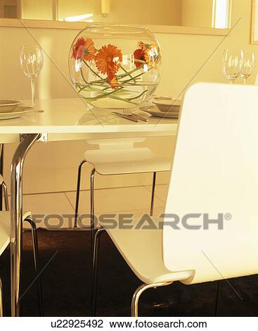 Stock Image - Close-up of orange gerberas in globe bowl on modern cream table with cream chair. Fotosearch