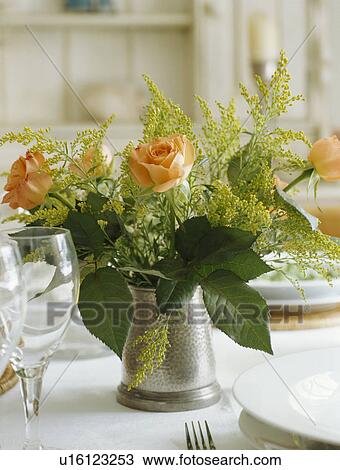 Stock Image - Close-up of orange roses in pewter jug on table. Fotosearch