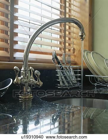 Stock Image - Close-up of stainless steel mixer tap pouring water into kitchen sink below window with slatted wooden blind. Fotosearch