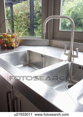 Stock Image - Close-up of stainless-steel mixer tap pouring water into stainless-steel double sink. Fotosearch