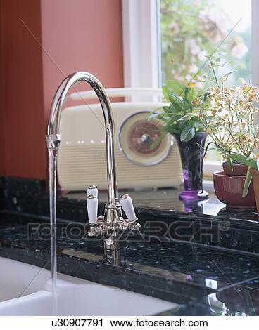 Stock Image - Close-up of stainless steel tap pouring water into sink below window sill with cream radio. Fotosearch