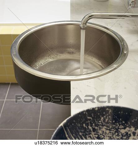 Stock Photograph - Close-up of steel tap pouring water into circular stainless steel sink. Fotosearch