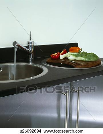 Picture - Close-up of tap pouring water into circular stainless steel sink in black worktop. Fotosearch