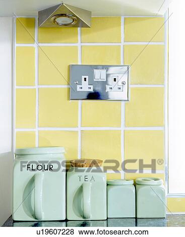 Stock Photo - Close-up of turquoise ceramic storage jars below electric plug socket on yellow tiled wall. Fotosearch