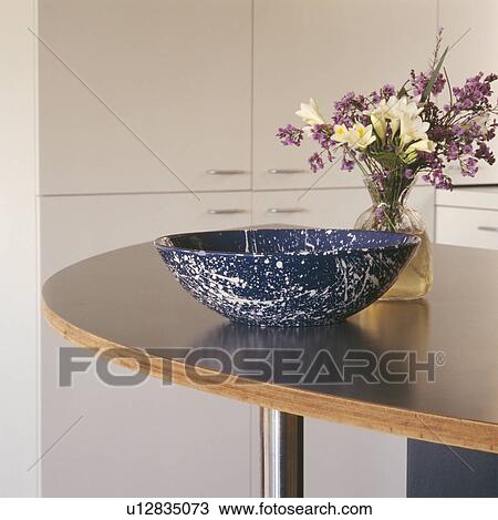Stock Image - Close-up of vase of flowers and large blue bowl on island unit. Fotosearch