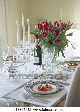 Stock Image - Close-up of vase of red tulips on table with white cloth and place settings for lunch. Fotosearch