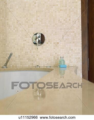 Stock Photograph - Close-up of wall-mounted tap in beige mosaic tiled wall above undermounted bath. Fotosearch