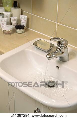 Picture - Close up of water pouring from chrome tap on white ceramic basin. Fotosearch