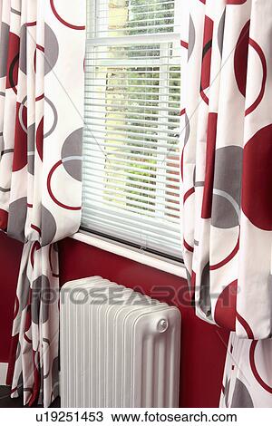 Stock Image - Close-up of white slatted blind and red patterned curtains on window above white radiator. Fotosearch