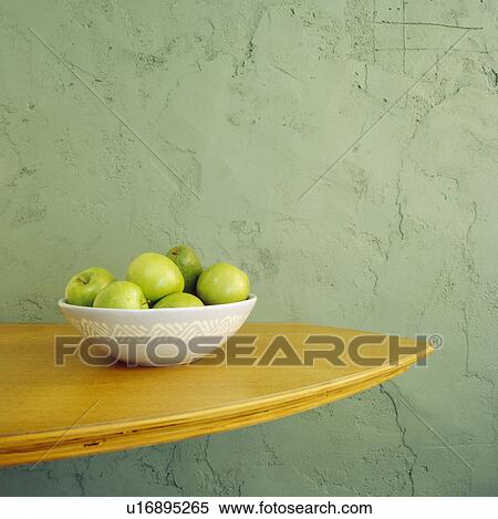 Stock Photography - Close-up up of green apples in bowl on wooden shelf on textured green wall. Fotosearch