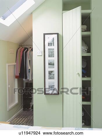 Clothes on hanging rail and cupboard door open on storage selves in pastel green attic conversion bedroom View Large Photo Image Picture - Clothes on hanging rail and cupboard door open on storage selves in pastel green attic conversion bedroom. Fotosearch